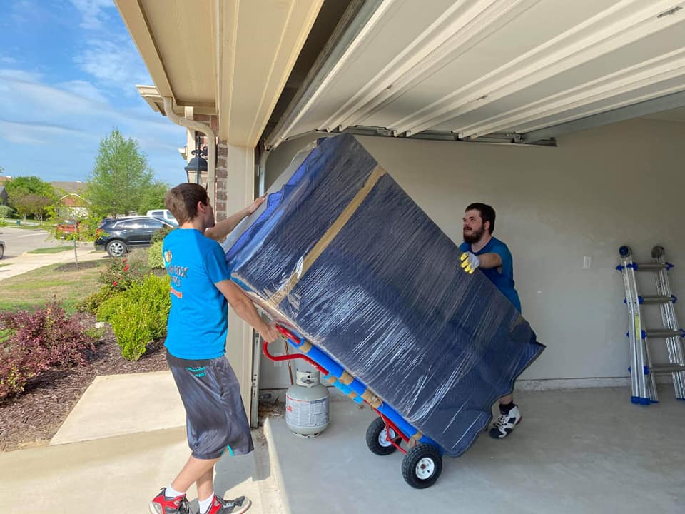 Movers wheeling a shrink-wrapped refrigerator out of a garage on a hand truck